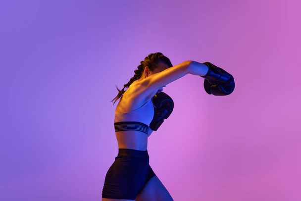 Athletisch und kraftvoll sticht die junge Frau in Boxerkleidung vor dem rosa-violetten Studiohintergrund in Neonlicht hervor. Konzept von Sport, aktivem Lebensstil, Gesundheit, Wettbewerb, Meisterschaft. - Foto, Bild