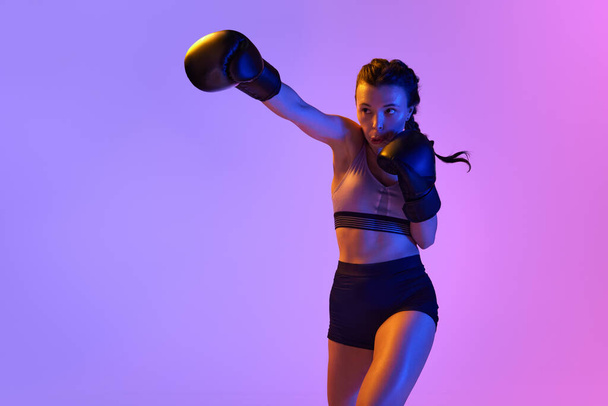 Intense moment as determined boxer girl meticulously training, preparing for combat against gradient studio background in neon light. Concept of sport, active lifestyle, health, competition. - Photo, Image