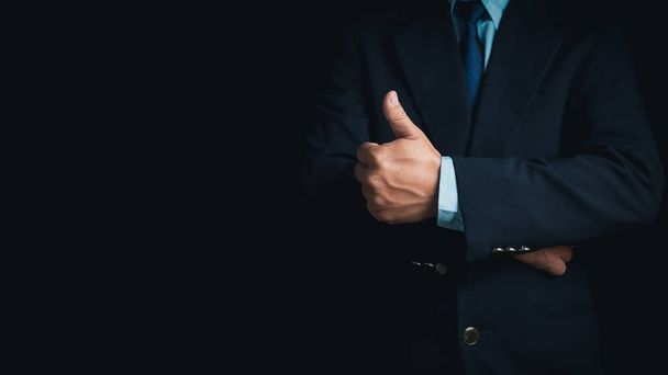 Un uomo d'affari in giacca e cravatta sta con le braccia incrociate e i pollici su uno sfondo scuro. - Foto, immagini