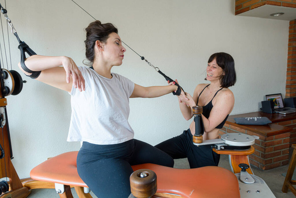Entrenador femenino caucásico de pie asistiendo a su estudiante durante el entrenamiento utilizando el ejercicio y la máquina de estiramiento - Foto, Imagen