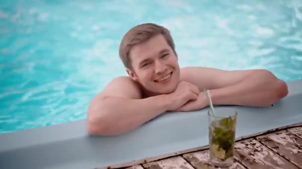 Shot of handsome happy man folded arms at swimming pool corner outdoors. Smiling boyfriend recreation in blue water enjoying his cold cocktail mojito. Satisfied guy on resort. High quality 4k footage - Footage, Video