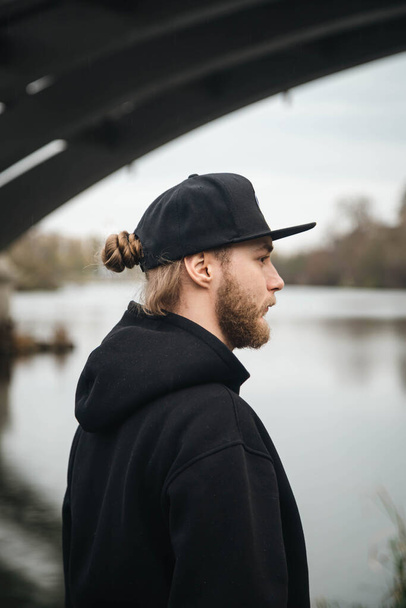 Ein stylischer junger bärtiger Mann mit Dutt, schwarzer Mütze und schwarzem Kapuzenpulli steht unter einer Brücke am Fluss und blickt in die Ferne.. - Foto, Bild