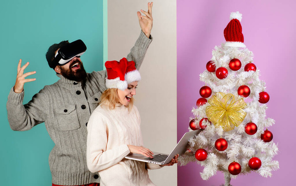 Wesołych Świąt w pobliżu choinki. Uśmiechnięta kobieta w kapeluszu Mikołaja z laptopem i szczęśliwym brodatym mężczyzną w okularach wirtualnej rzeczywistości na Boże Narodzenie. Technologia 3D i rozrywka. Wirtualna maska rzeczywistości - Zdjęcie, obraz
