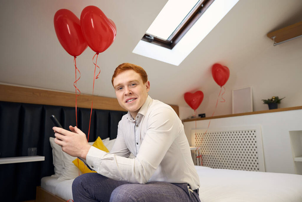 Maschio sorridente in camicia bianca seduto sul letto con palloncini cuore rosso e telefono in mano in camera da letto accogliente - Foto, immagini