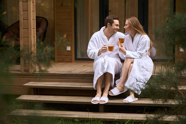Молодая пара в белых халатах пьет горячий чай во дворе и дышит свежим лесным воздухом - Фото, изображение
