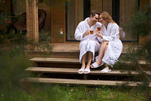 白いバスローブでかわいいカップルは,裏庭のラウンジと新鮮な森の空気を呼吸しながらお茶を飲んで - 写真・画像