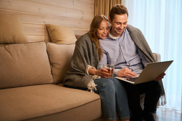 Bella coppia guardando lo schermo durante l'utilizzo di laptop seduto rilassato sul divano sotto plaid e bere tè - Foto, immagini