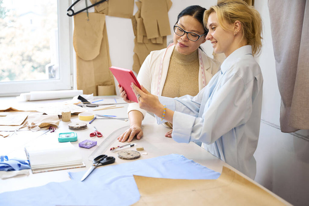Designers de moda feminina usam um tablet em um ambiente de trabalho, há um monte de suprimentos de costura na mesa - Foto, Imagem