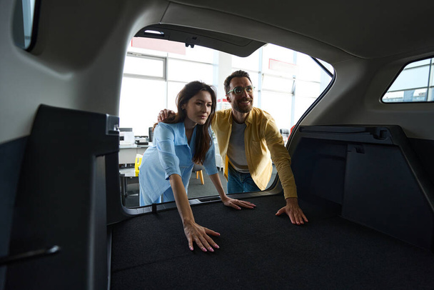 Gözlüklü çalışkan bir adam ve şık elbiseli çekici bir kadın araba galerisindeki araba bagajını kontrol ediyor. - Fotoğraf, Görsel