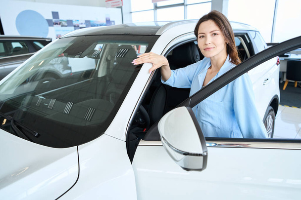 Aantrekkelijke vrouw in stijlvolle kleding op zoek naar camera tijdens het openen van de auto deur in de auto showroom - Foto, afbeelding