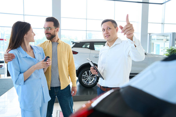 Happy Caucasian woman and diligent man looking at each other while consultant talking to them in car dealership - Photo, Image