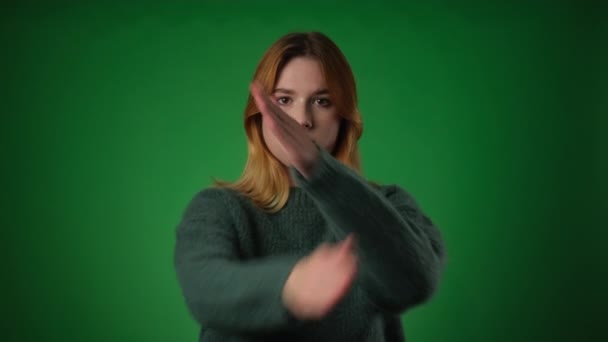 Determined Caucasian woman, dressed casually, crosses arms in a stop gesture against a vibrant green backdrop. Expressions and the crossed arms convey a sense of firmness and a clear signal to halt. - Footage, Video