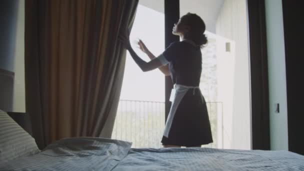 Medium wide shot of young room attendant opening and adjusting curtains in hotel room, preparing it for new guests - Footage, Video