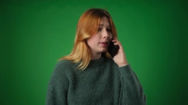 Distraught woman, dressed casually, engages in a phone conversation with an upset expression against a green backdrop. Expressions of distress and the phone call convey a sense of emotional turmoil. - Footage, Video