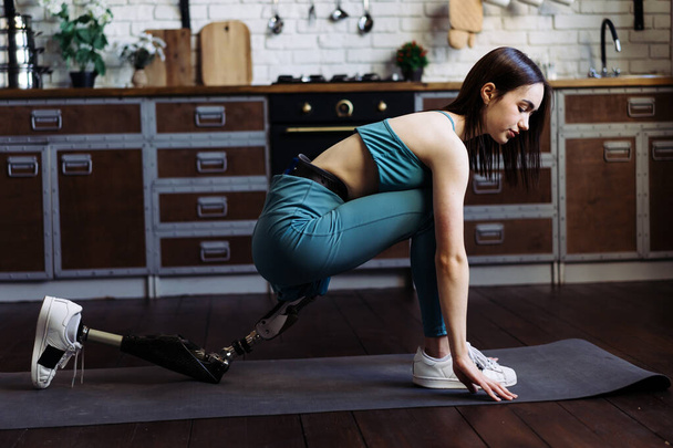 Female with leg prosthesis balances in yoga position on mat at home. Lady performs exercises on floor of kitchen demonstrating balance and strength - Photo, Image