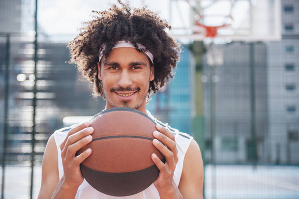 Atractivo jugador de baloncesto está sosteniendo una pelota, mirando a la cámara y sonriendo mientras está de pie en la cancha de baloncesto al aire libre - Foto, imagen