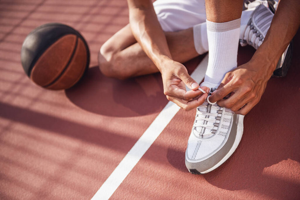 Bijgesneden afbeelding van basketballer vetersluiting zijn trainers zittend op het basketbalveld buitenshuis - Foto, afbeelding