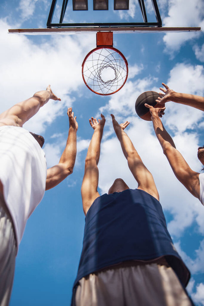 Нижний вид красивых баскетболистов, стреляющих мячом через обруч во время игры на баскетбольной площадке на открытом воздухе
 - Фото, изображение