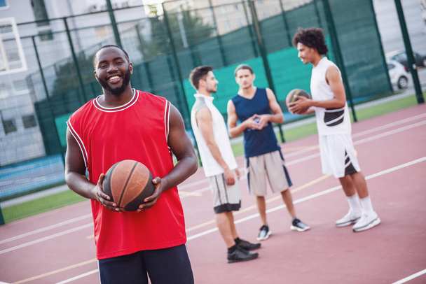 Красивые баскетболисты разговаривают и улыбаются на баскетбольной площадке на открытом воздухе, парень на переднем плане смотрит в камеру
 - Фото, изображение
