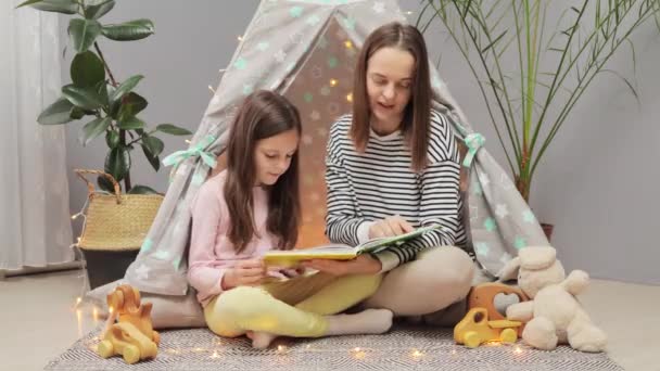 Brown haired woman with her little daughter reading book in hovel at home doing home work preparing for literature lesson together while sitting on floor. - Video, Çekim
