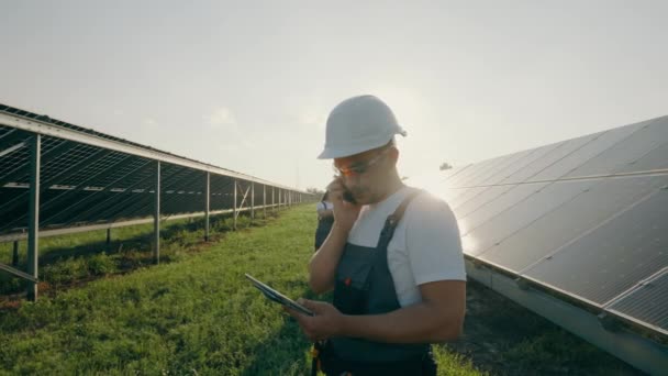 Egy rendező mérnök portréja, amint mobilon beszél, miközben egy naperőműben áll. Egy munkás, aki napelemeket vizsgál a háttérben. Kiváló minőségű 4K felvételek - Felvétel, videó
