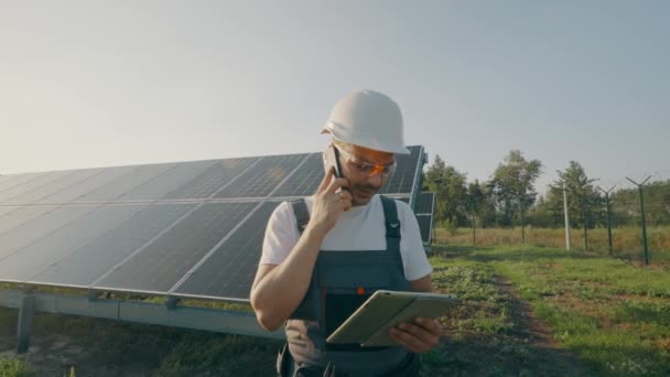 Bir güneş çiftliğinde dururken cep telefonuyla konuşan bir mühendisin portresi. Arka plandaki güneş panellerini inceleyen bir işçi. Yüksek kalite 4K görüntü - Video, Çekim
