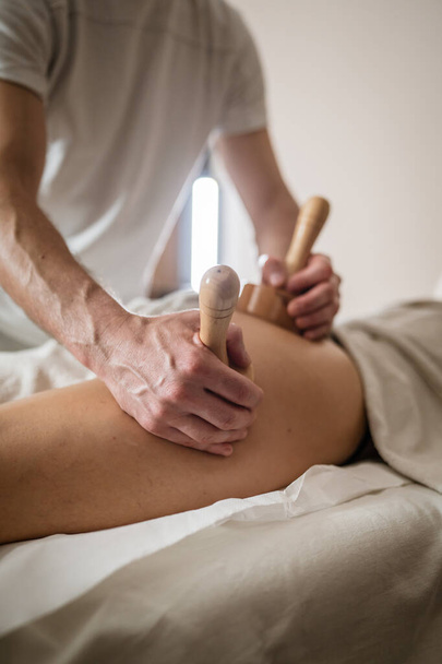Unknown Caucasian woman having madero therapy massage anti-cellulite treatment by professional therapist holding wooden tools in hands in studio or salon with copy space - Photo, Image