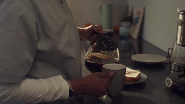Closeup kantelen schot van Afro-Amerikaanse verpleegster in medische uniform staan in de keuken thuis, het gieten van koffie uit kan in mok, het nemen van sandwich van bord en eten - Video