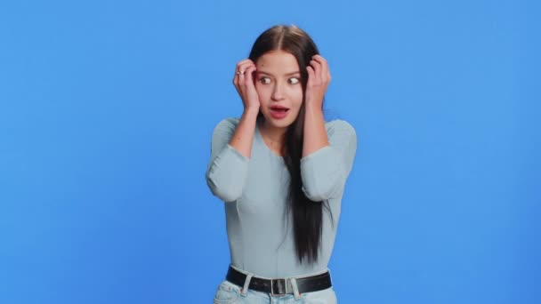 Excited amazed woman touching head and showing explosion, looking worried and shocked, celebrating success idea. Looking surprised of win wow brunette girl isolated alone on blue studio background - Footage, Video