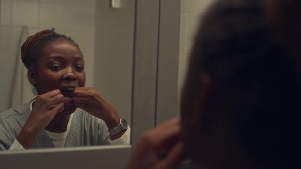 Крупным планом через плечо снята молодая черная медицинская работница с плетеными волосами в булочке, в медицинской форме, стоящая в ванной перед зеркалом, зубная нить, готовясь к работе - Кадры, видео