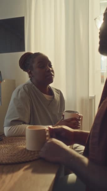 Plan vertical moyen avec éclat de lentille de jeune femme afro-américaine en uniforme d'infirmière assise dans la cuisine à la maison avec son mari, buvant du thé, bavardant et discutant des problèmes de travail - Séquence, vidéo