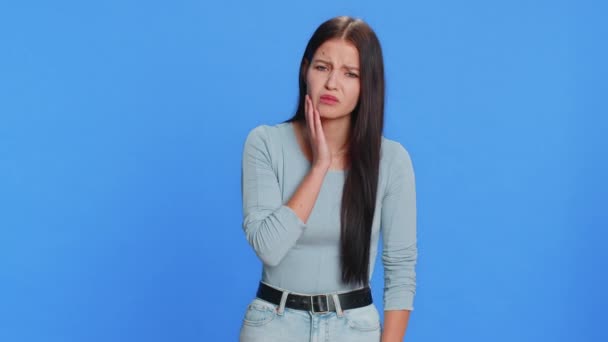 Dental problems. Young woman touching cheek, closing eyes with expression of terrible suffer from painful toothache, sensitive teeth, cavities. Pretty brunette girl isolated on blue studio background - Footage, Video