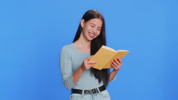 Happy relaxed smiling Caucasian young woman reading funny interesting fairytale book, leisure hobby, knowledge wisdom, education, learning, study, wow. Девушка-подросток изолирована на синем фоне - Кадры, видео