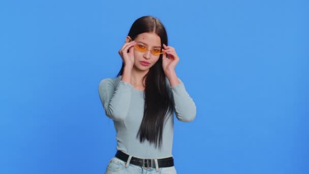 Caucasian young stylish cool woman wearing yellow sunglasses, charming friendly playful smile, celebrate victory win. Teenager happy girl feel satisfied excitement isolated on blue background indoors - Footage, Video