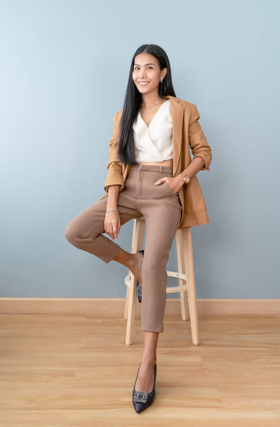 Retrato de sonreír a una atractiva mujer de negocios asiática guerreando traje marrón fresco chic mientras está sentado en la silla y mirando a la cámara aislado fondo verde pastel. Longitud completa, espacio de copia. - Foto, imagen