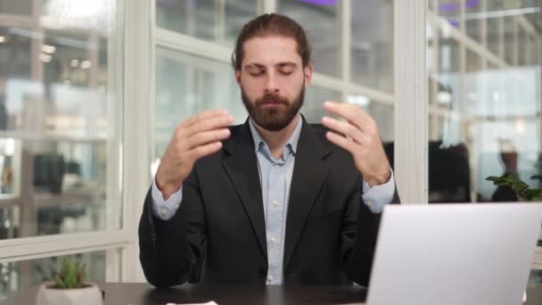 Homme d'affaires barbu assis à table devant un ordinateur portable moderne et les doigts pliants dans le geste mudra tout en méditant à l'intérieur. Caucasien pigiste fermer les yeux et se détendre pendant la pause sur le lieu de travail. - Séquence, vidéo