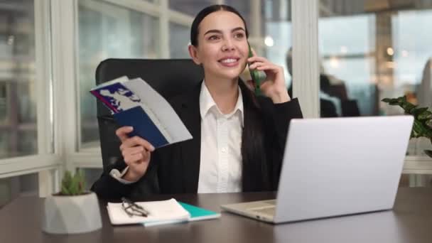 Smiling manager sitting at workplace in office with digital laptop and positively discussing vacation on smartphone. Caucasian female keeping foreign passport and tickets while planning new journey. - Footage, Video