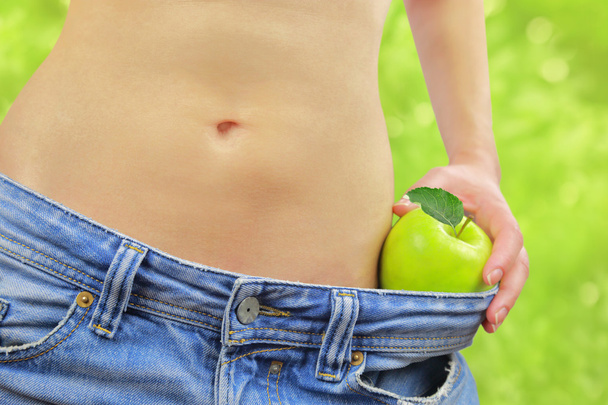 Frauenbauch mit Apfel und übergroßen Jeans, auf grünem Bokeh-Hintergrund - Foto, Bild
