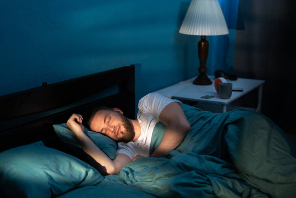 Красивый кавказский мужчина спит уютно на кровати в своей спальне по ночам с голубыми ночными красками в комнате. Копирование пространства - Фото, изображение