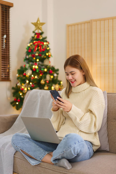 冬の季節のコンセプト, 女性は,ラップトップに取り組んでおり,クリスマスツリーの近くの電話でオンラインショッピング. - 写真・画像