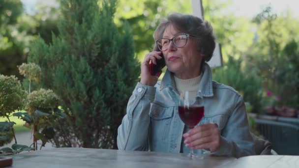 Gelukkig leven van succesvolle oude dame in denim shirt met een zakelijk telefoontje tijdens het drinken van alcohol op het terras. Positieve oudere vrouw drinkt wijn tijdens een buitenlandse reis. Rode wijn en een gesprek. Hoog - Video