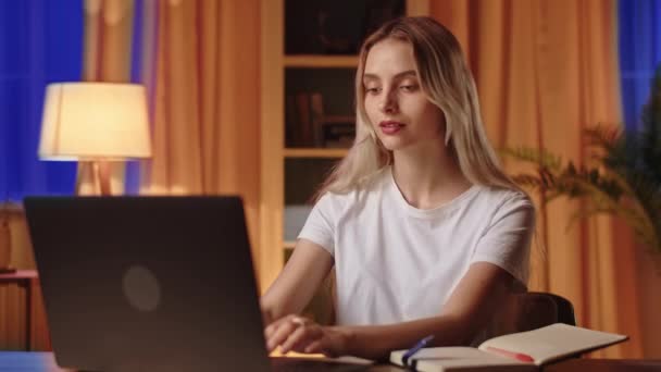 Блондинка студентка використовує ноутбук, шукає інформацію, щоб підготуватися до занять або тез і записує нотатки в блокноті. Молода леді вчителька спілкується з школярами віддалено через - Кадри, відео