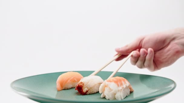Nahaufnahme einer männlichen Hand, die mit Essstäbchen ein Aal-Sushi von einem Teller mit Lachs und Shrimp-Sushi auf weißem Hintergrund nimmt - Filmmaterial, Video