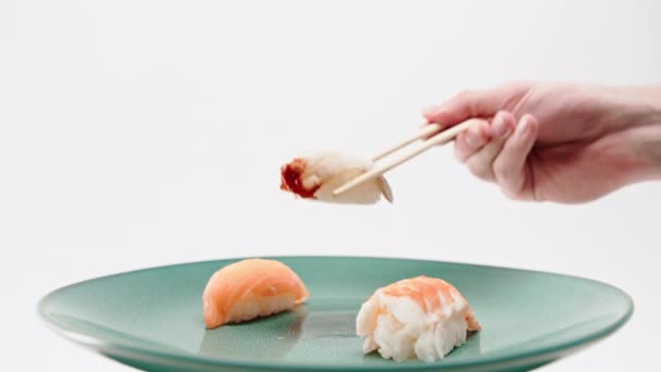 Nahaufnahme einer männlichen Hand, die mit Essstäbchen ein Aal-Sushi von einem Teller mit Lachs und Shrimp-Sushi auf weißem Hintergrund nimmt - Filmmaterial, Video