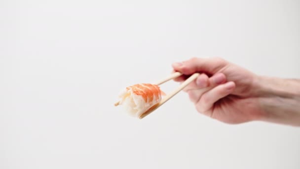 白い背景に空中にエビの寿司のナイリを保持する箸を持つ男の手のクローズアップ - 映像、動画