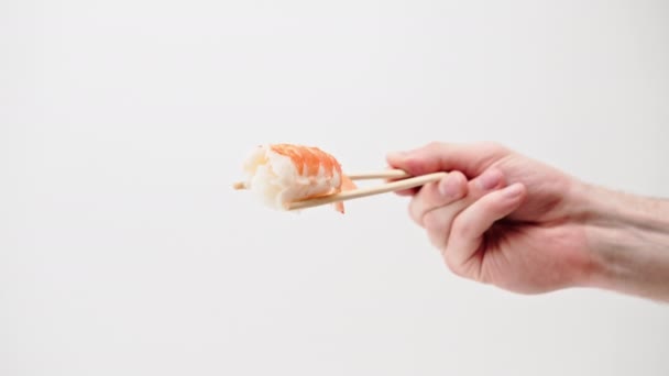 白い背景に空中にエビの寿司のナイリを保持する箸を持つ男の手のクローズアップ - 映像、動画
