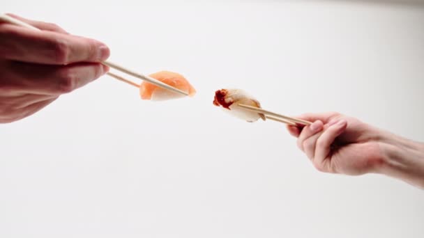 Közelkép férfi kezekről evőpálcikákkal, amint angolnás sushit és lazacszusit tartanak egymás előtt, fehér alapon. - Felvétel, videó