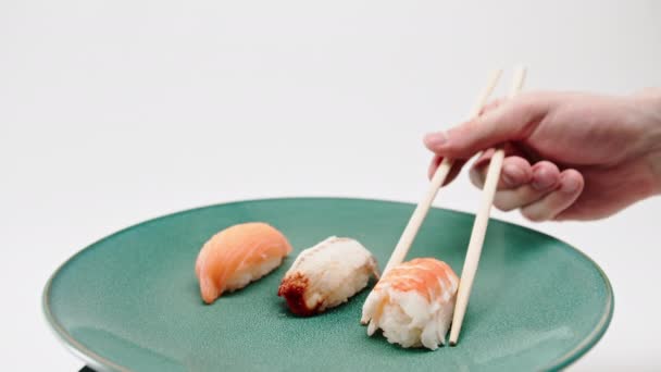Крупный план мужской руки с помощью палочек для еды, чтобы взять один суши креветок из тарелки лосося и угрей суши на белом фоне - Кадры, видео