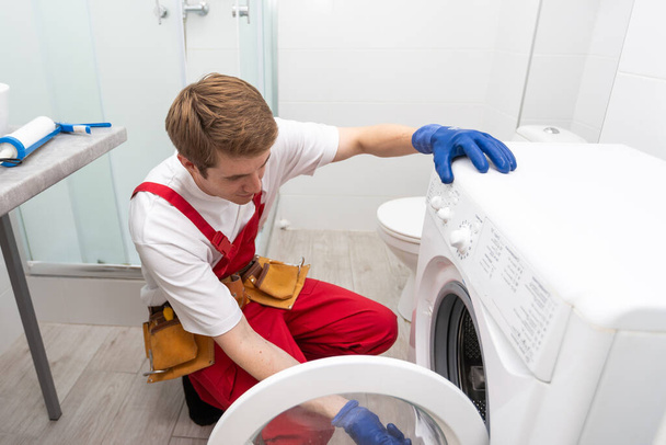 Le jeune beau réparateur en costume de travailleur avec la boîte à outils professionnelle fixe la machine à laver dans la salle de bain - Photo, image
