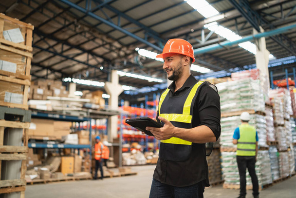 Professional manager man employee smile using tablet check stock working at warehouse. Worker wearing high visibility clothing and a hard hat, helmet and checking and count up goods boxes delivery. - Photo, Image